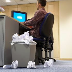 Man sitting at desk with trashcan piled up and crumpled paper on the floor