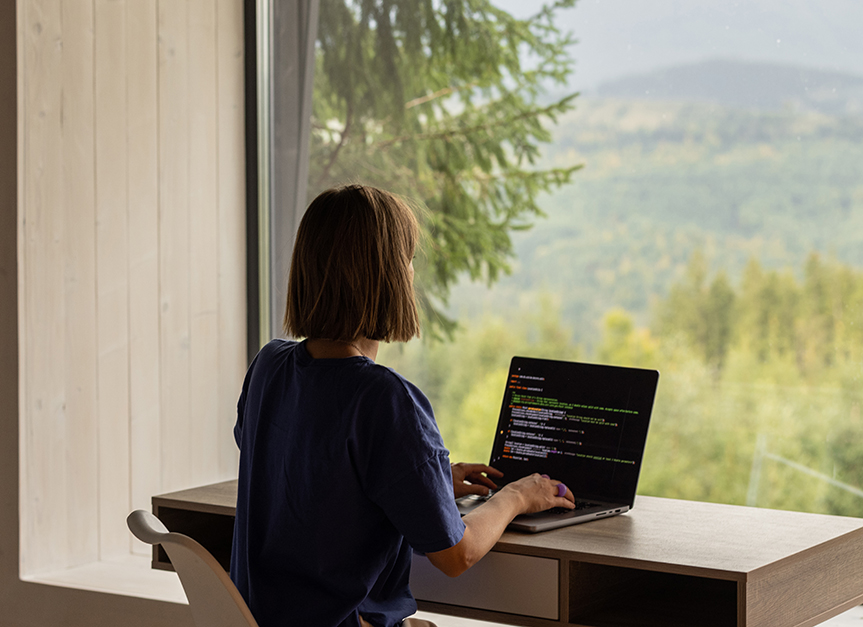 Image of a woman with her back to the camera working on a laptop facing a window with a nature scene outside